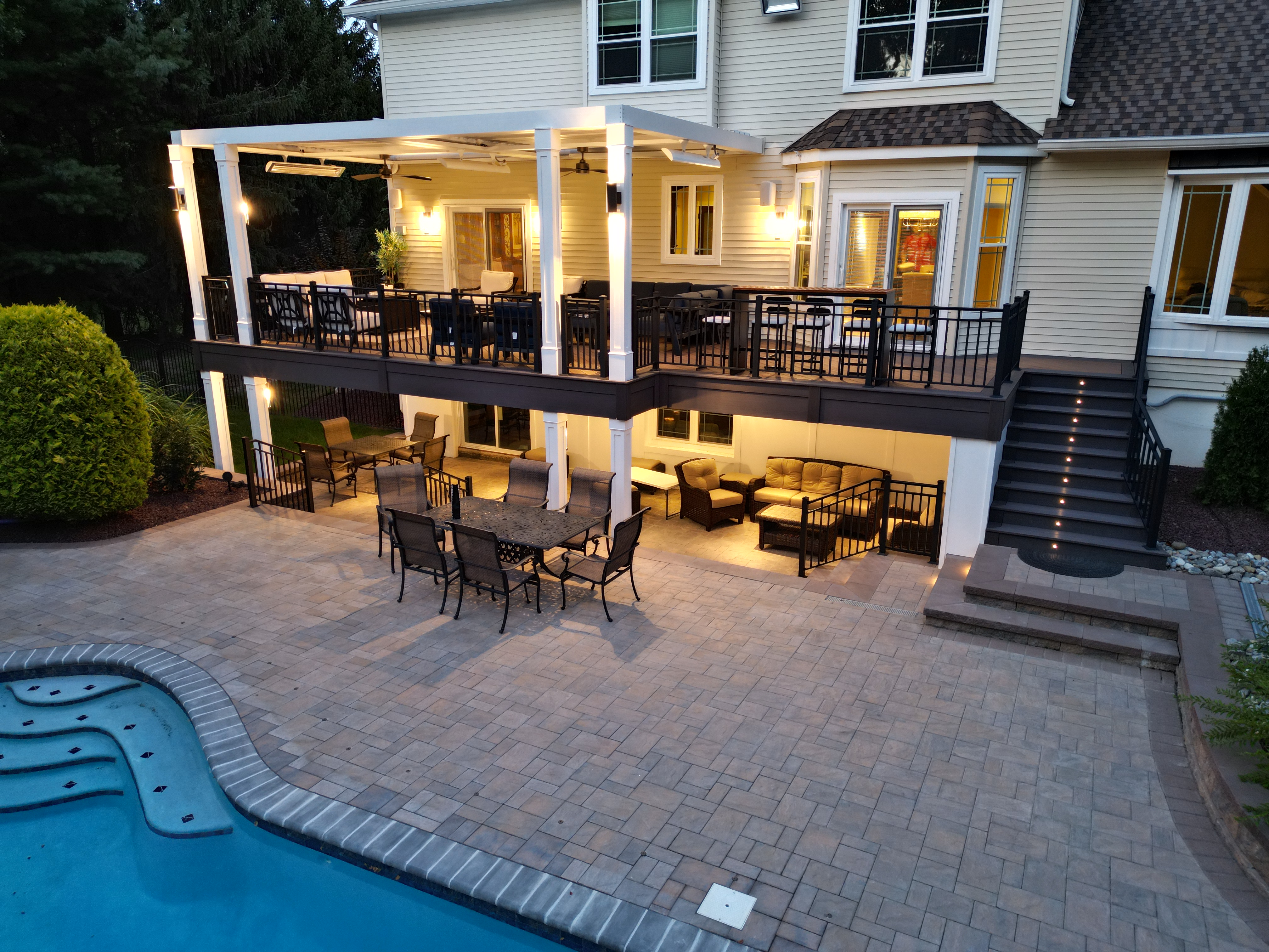 finished under deck patio with multiple seating areas, outdoor dining set, and poolside access beneath an upper deck with lighting and covered lounge