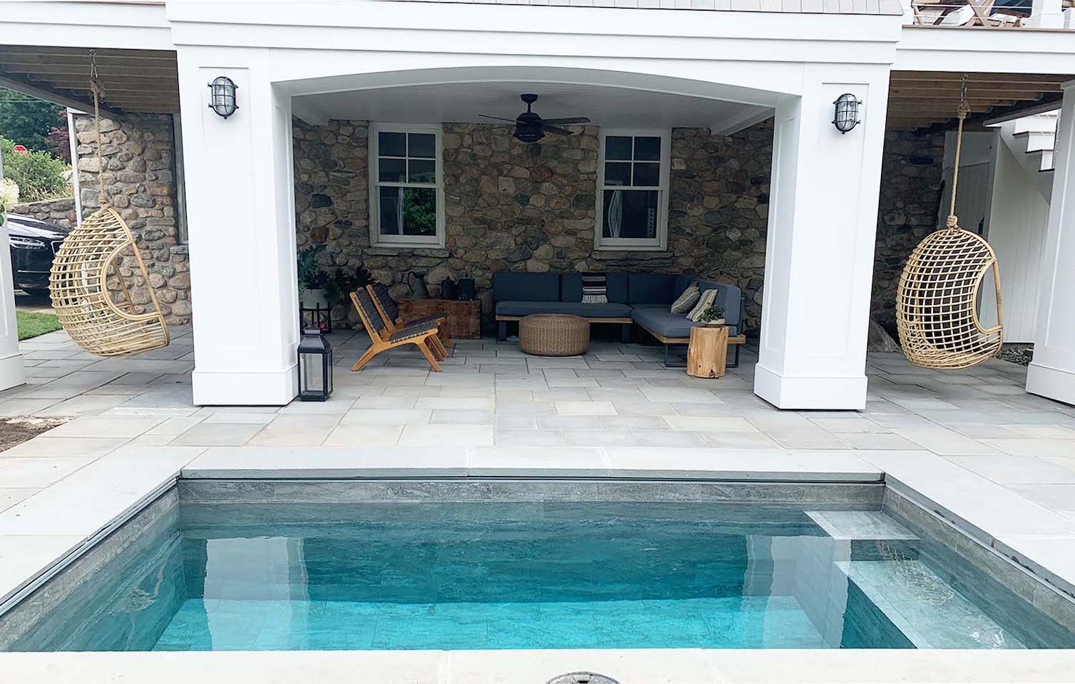 small pool beside covered patio lounge with stone wall backdrop, hanging chairs, and outdoor seating area