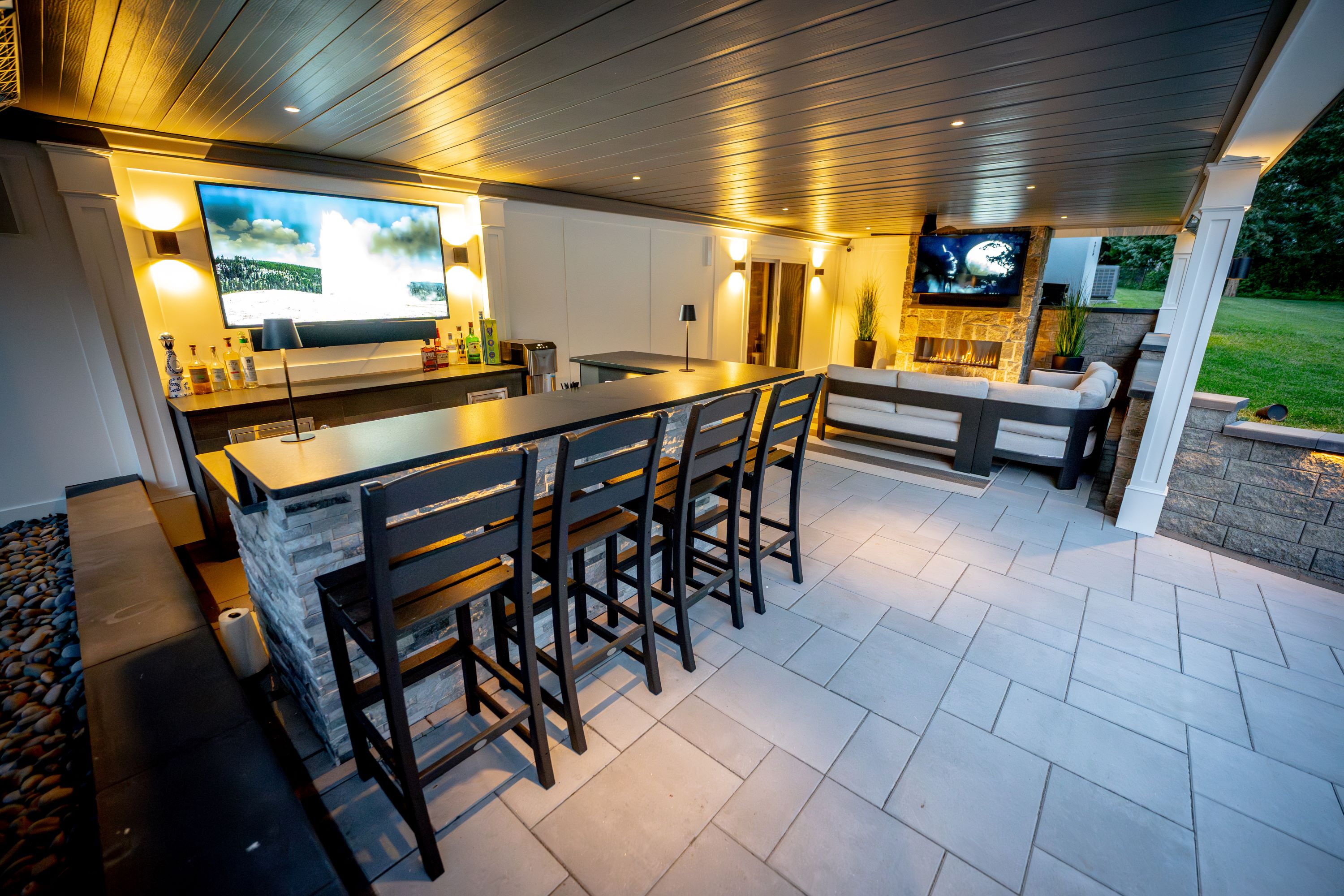 finished under deck entertainment space with built-in bar, stacked stone base, bar seating, large wall-mounted TV, recessed lighting, lounge area with stone fireplace, and paver flooring